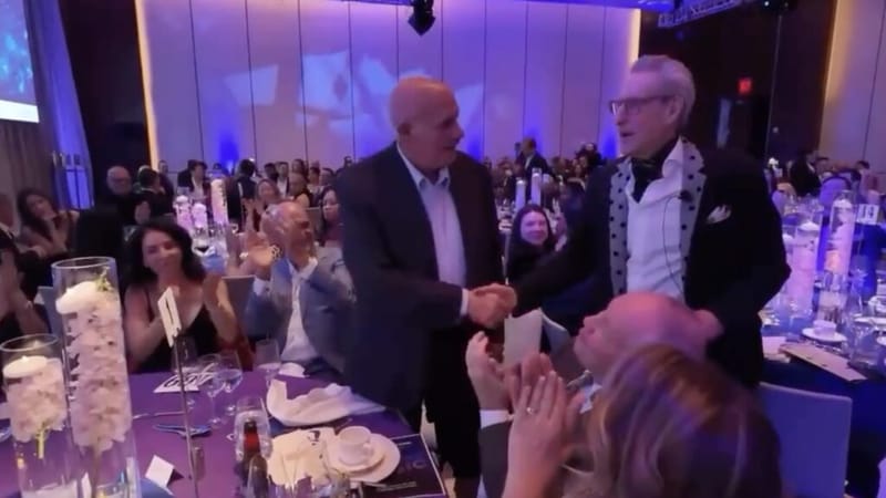 Gene Seidman shaking hands with a guest at a charity gala, surrounded by applauding attendees at candlelit tables