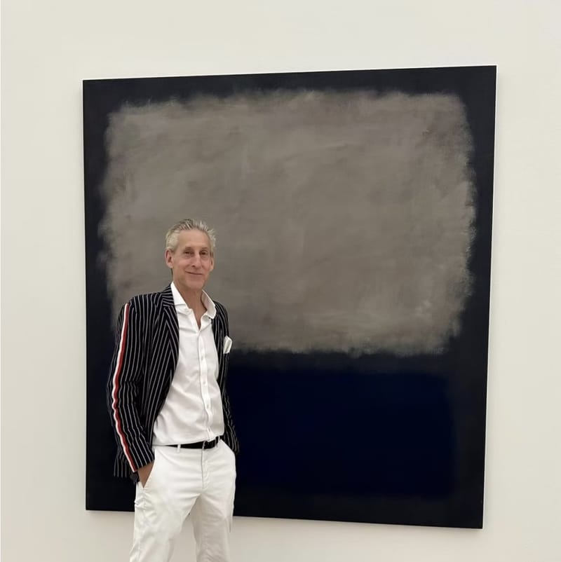 Gene Seidman standing in front of a large dark abstract painting, wearing a pinstripe blazer and white trousers