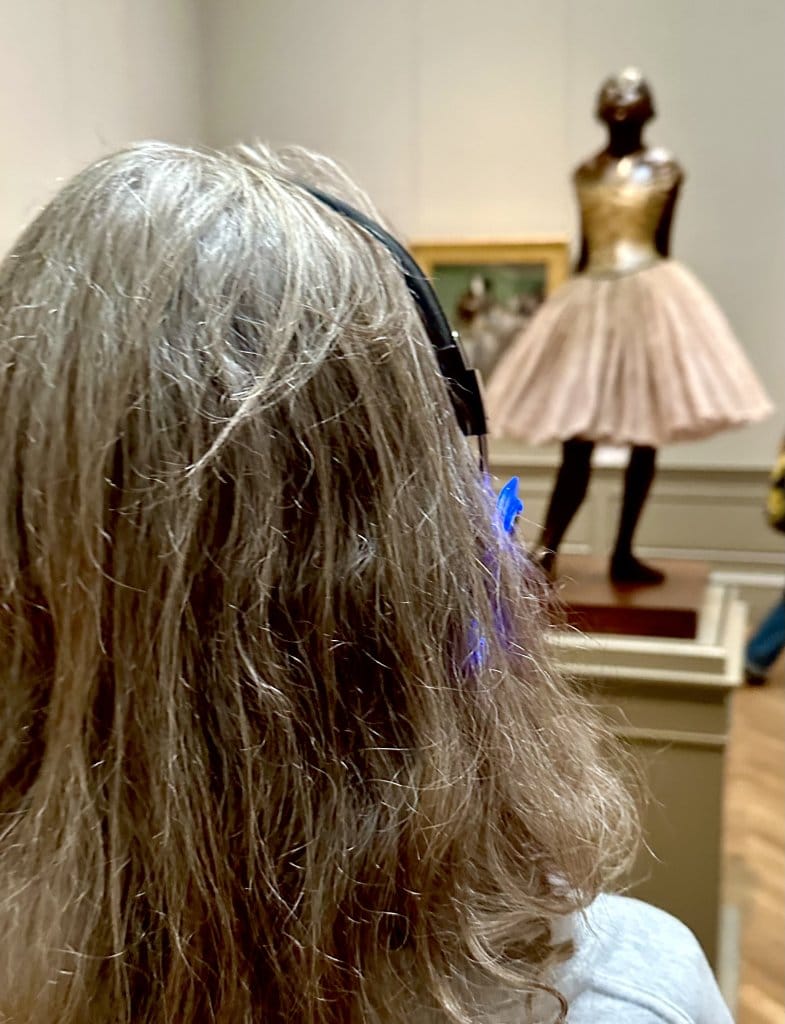 Museum visitor seen from behind, wearing headphones, looking at Degas' Little Dancer sculpture in a gallery